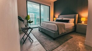 A modern bedroom with a bed, bedside tables, a plant, and a view of a cityscape through large windows. - Smart Brickell Hotel (Miami)