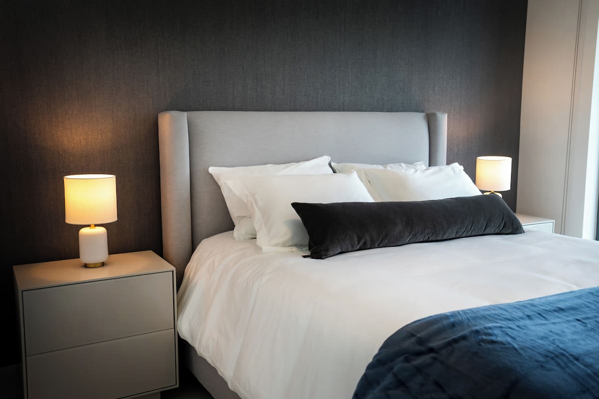A neatly made bed with white and dark pillows, a grey headboard, and two bedside lamps on a nightstand.