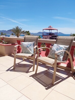 Two cushioned chairs with patterned cushions on a tiled terrace overlooking a scenic view of the sea and mountains.