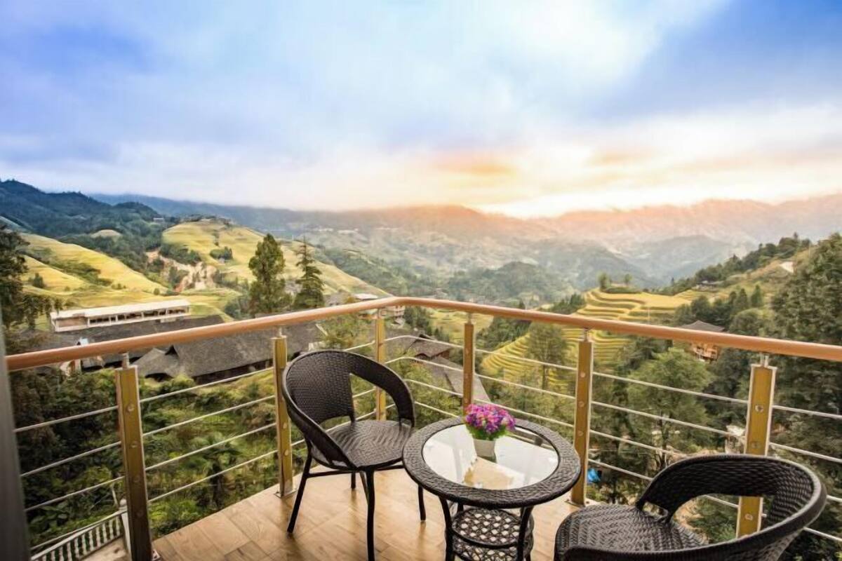 Un balcon agrémenté de fauteuils et d’une table en osier, d’où l’on peut admirer des champs en terrasses et des collines lointaines.