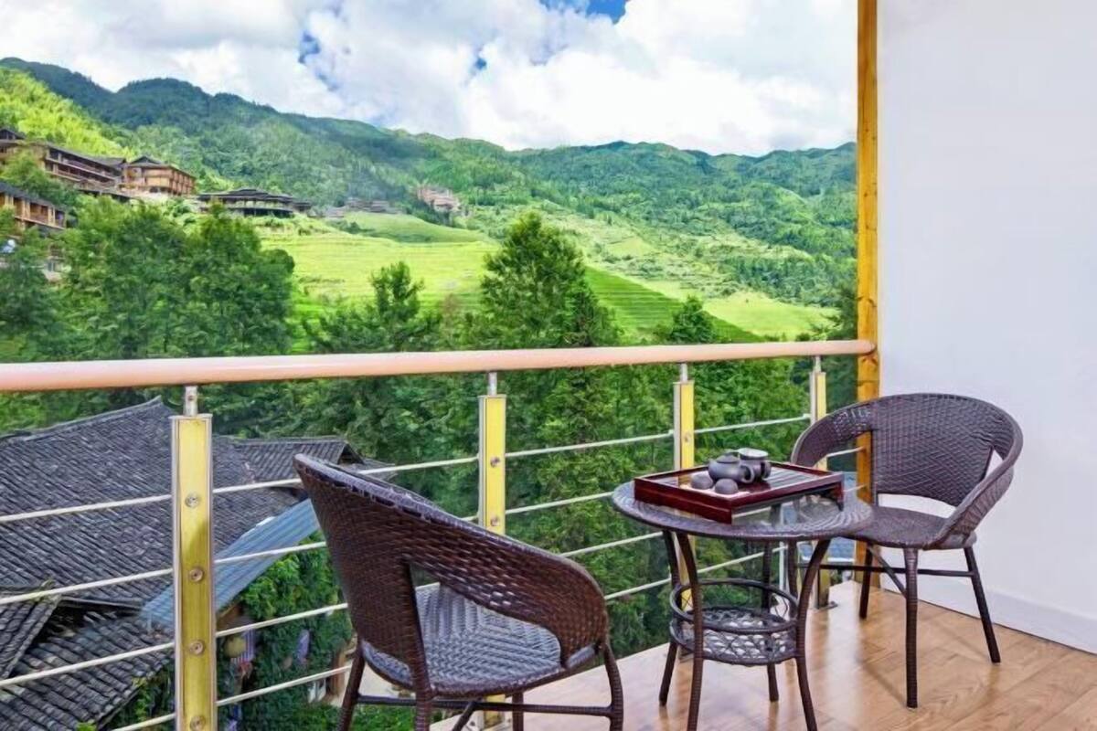 Un balcon doté de fauteuils et d’une table en osier, offrant une vue sur un paysage montagneux parsemé de bâtiments traditionnels.