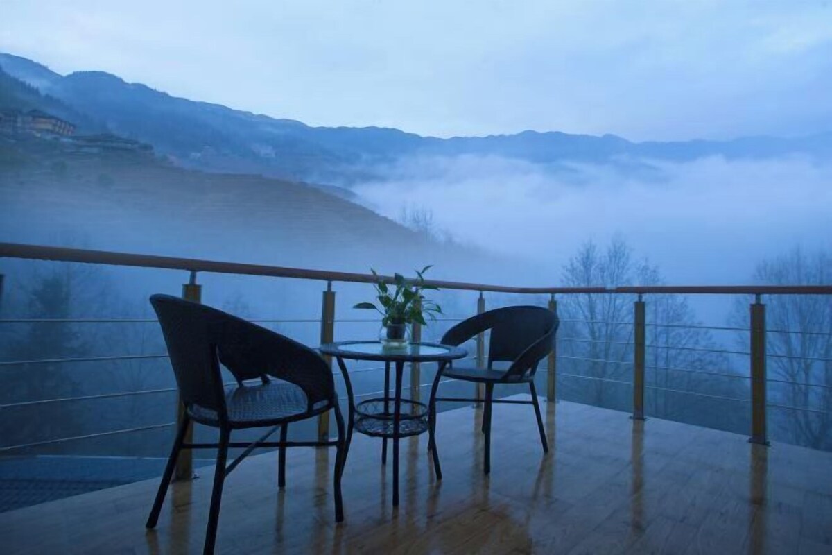 A balcony with a table and chairs, overlooking a mountainous landscape with fog.