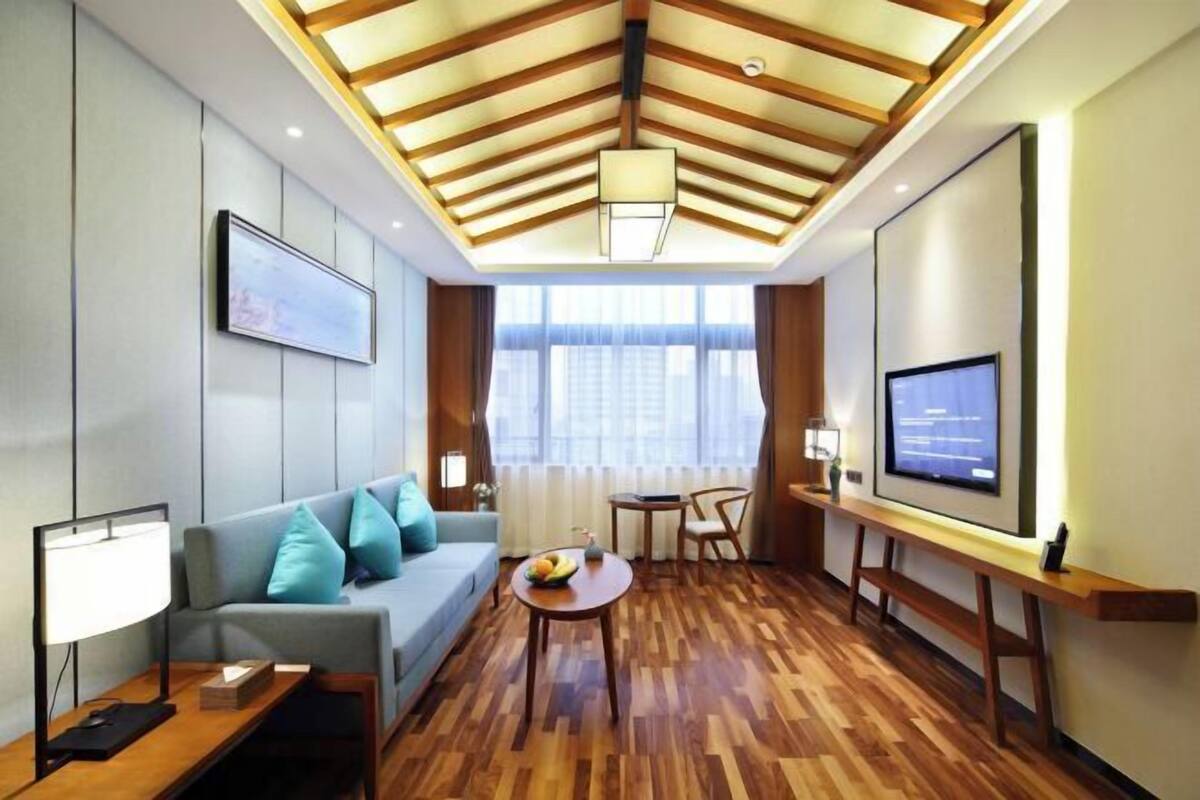 A modern hotel room with a wooden ceiling, a sofa with cushions, a small round table, a TV, and a window with curtains.
