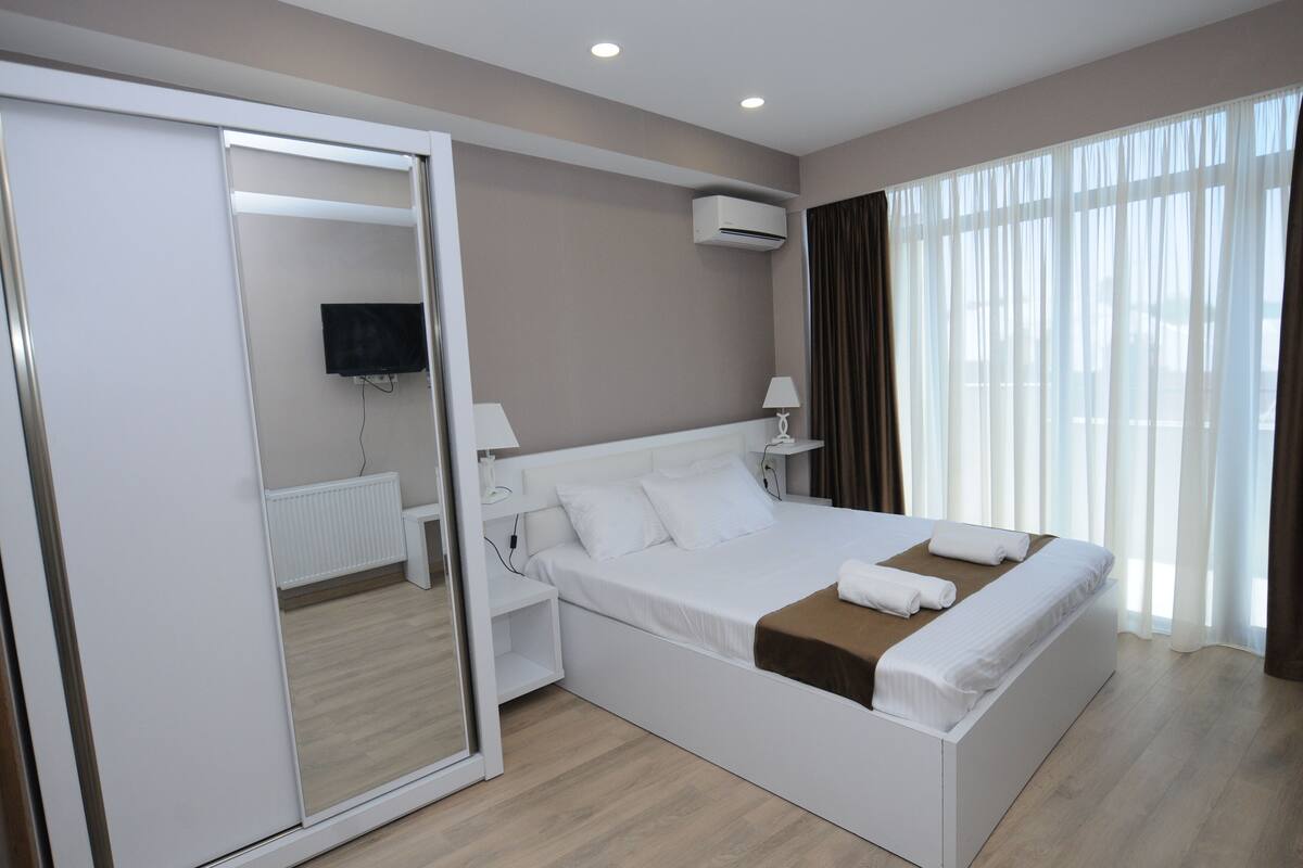 A modern hotel room with a large bed, a flat-screen TV, a bedside table, and a window with curtains.