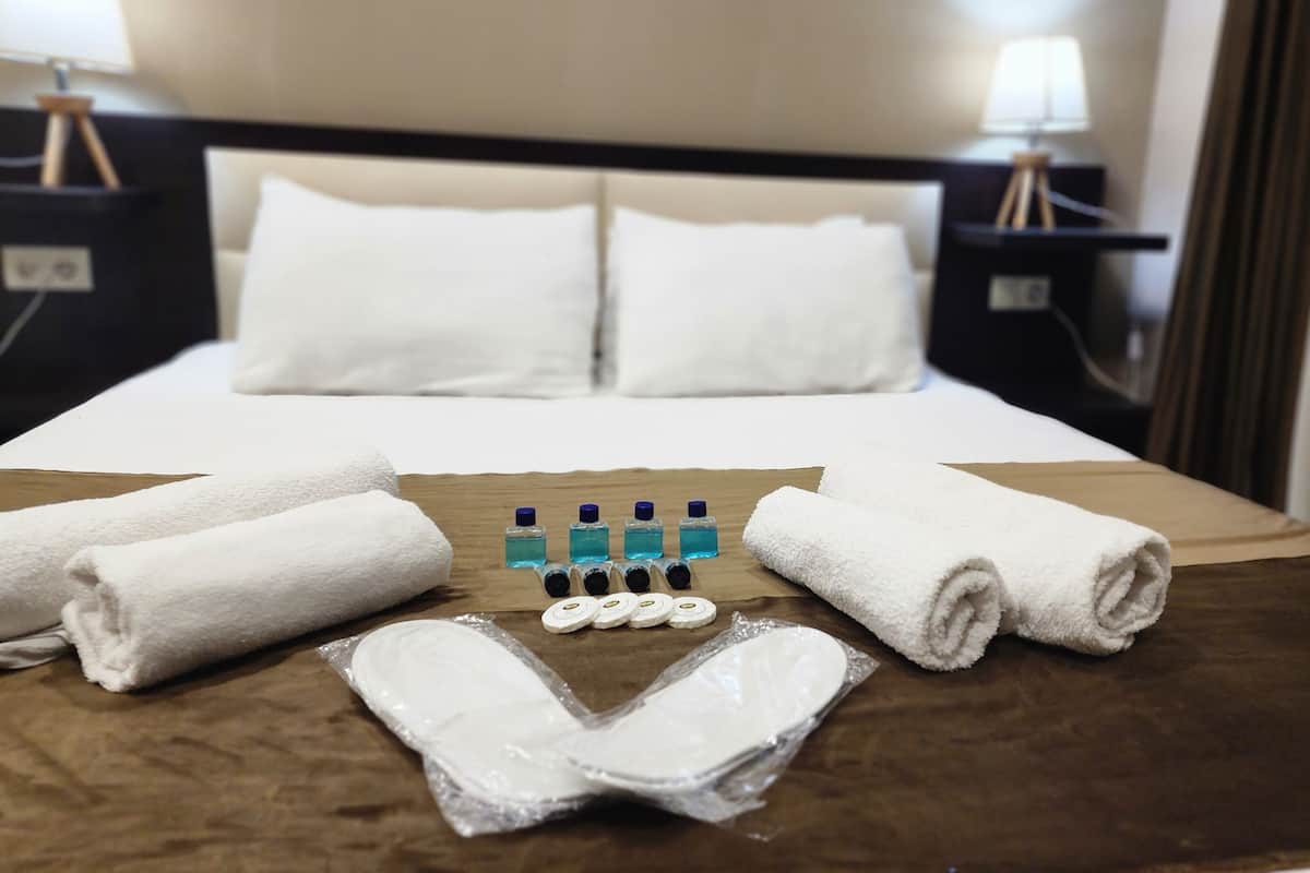 A neatly made bed with white linens, a brown bedspread, and a set of toiletries including a toothbrush, toothpaste, and a small bottle of liquid soap.