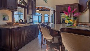 Una cocina amplia con armarios de madera oscura, una isla grande con taburetes de bar y vistas al océano a través de las ventanas.