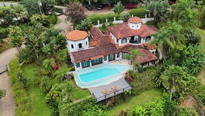 Una casa grande de varios pisos con piscina, rodeada de exuberante vegetación y palmeras.