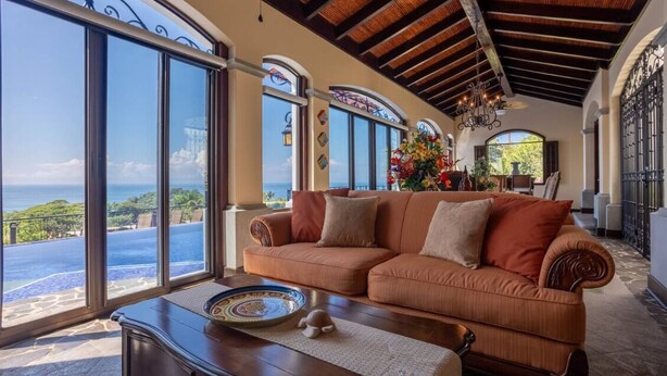 Amplia sala de estar con un gran ventanal que ofrece vistas panorámicas al océano, un sofá naranja acogedor y una mesa decorativa.