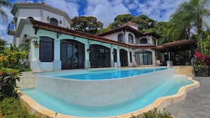 Una piscina amplia con borde de azulejos, rodeada de un jardín ajardinado y un edificio de dos plantas con ventanas de arco y balcones.