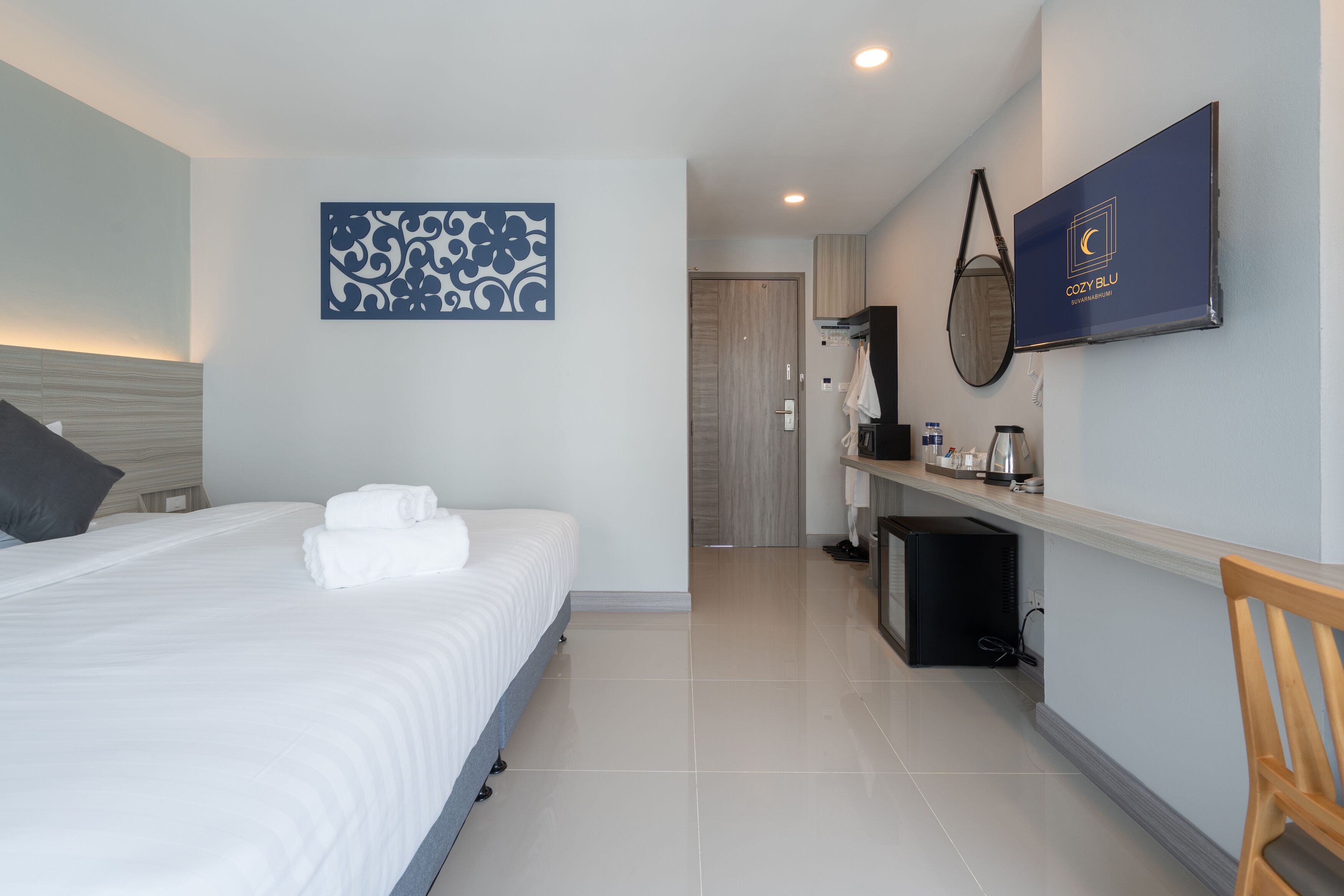 Une chambre d’hôtel moderne avec un lit bien fait, un téléviseur à écran plat fixé au mur, un petit coin salon avec un fauteuil et une porte donnant accès à la salle de bain.