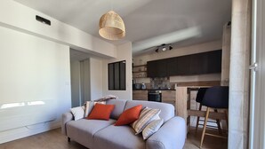 Una sala de estar moderna con un sofá gris, una lámpara colgante de fibra y una cocina al fondo.
