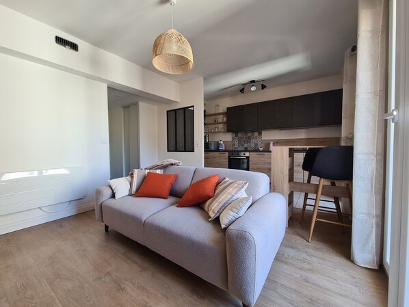 Una sala de estar moderna con un sofá gris, una lámpara colgante de fibra y una cocina al fondo.