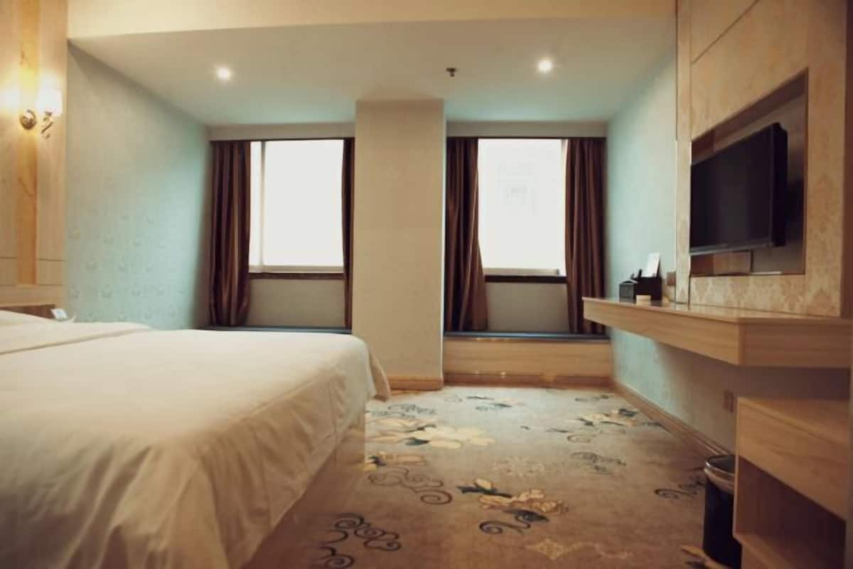 A hotel room with a bed, two windows with curtains, a television on a wooden shelf, and a patterned carpet.