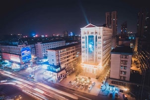 Un paisaje urbano nocturno con edificios iluminados, un edificio central destacado con techo rojo, y una intersección concurrida con estelas de luz de vehículos en movimiento.