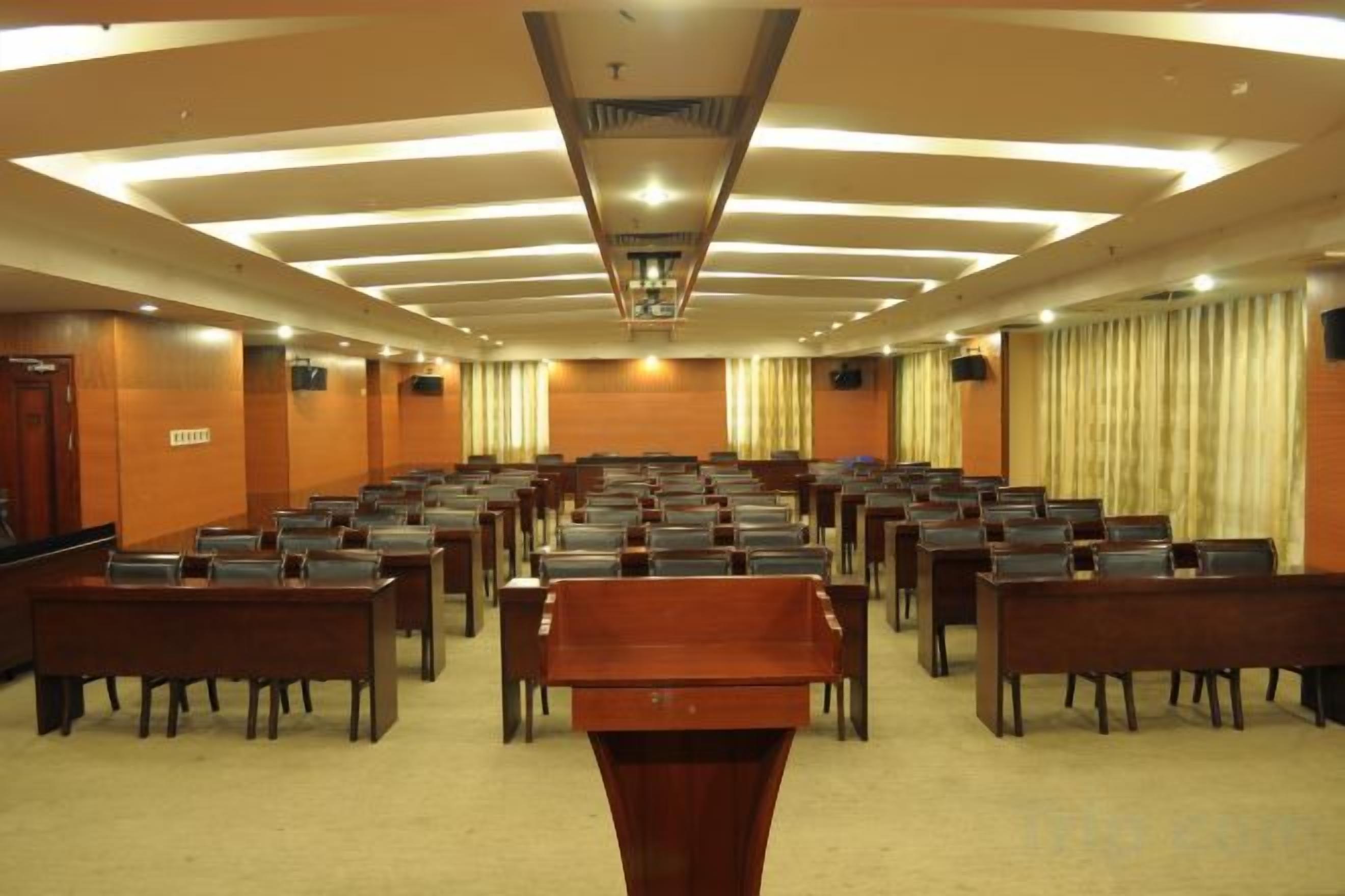 Un salón de conferencias con filas de mesas y sillas, un atril y un ventanal con cortinas.
