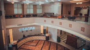 Un hall de hotel con un mostrador de recepción curvo, una zona de estar más baja y una segunda planta con mesas y sillas.