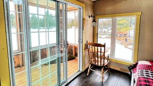 Una habitación con un gran ventanal corredero que da a una terraza exterior, una silla de madera, una mesita y una cama con manta a cuadros.
