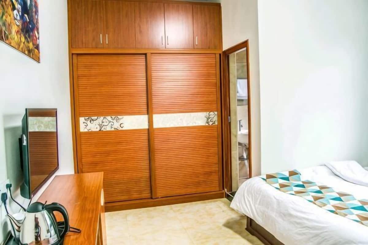 A bedroom with a wooden wardrobe, a bed with patterned bedding, a desk with a mirror, and a television.