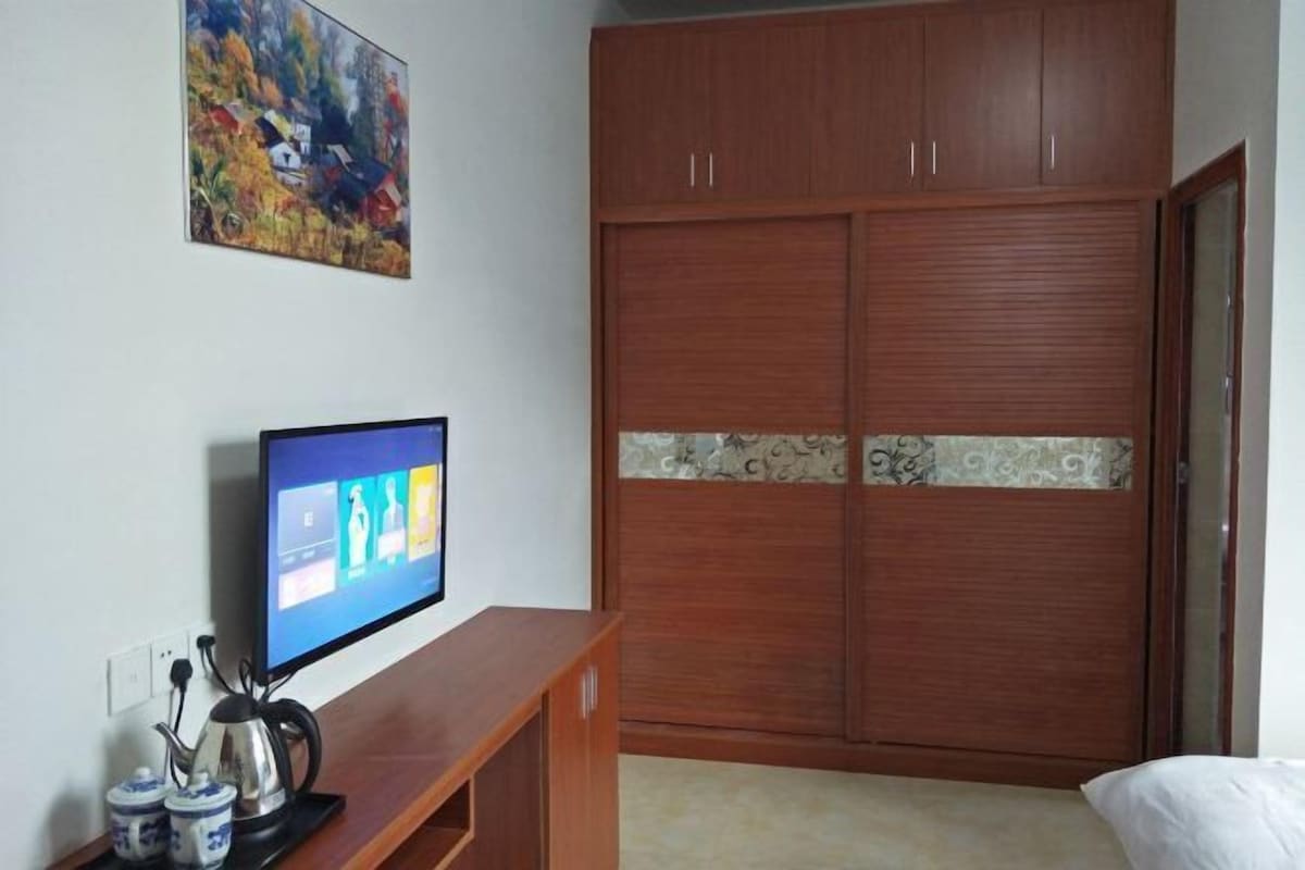 A bedroom with a wooden wardrobe, a television on a wooden stand, and a painting on the wall.