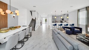 Eine moderne Küche mit Kochinsel, ein Essbereich mit langem Tisch und Stühlen sowie eine Wohnbereich mit Sofa und blauen Sesseln.