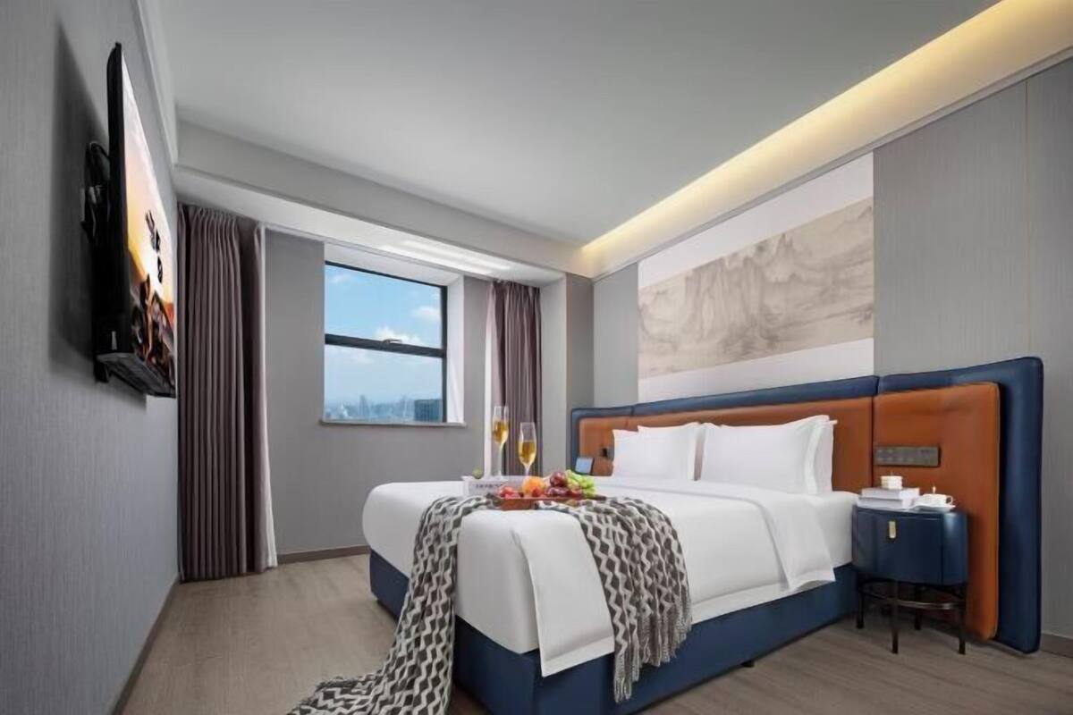 Una habitación de hotel moderna con una cama grande, un televisor de pantalla plana montado en la pared, una ventana con cortinas y vistas a la ciudad.