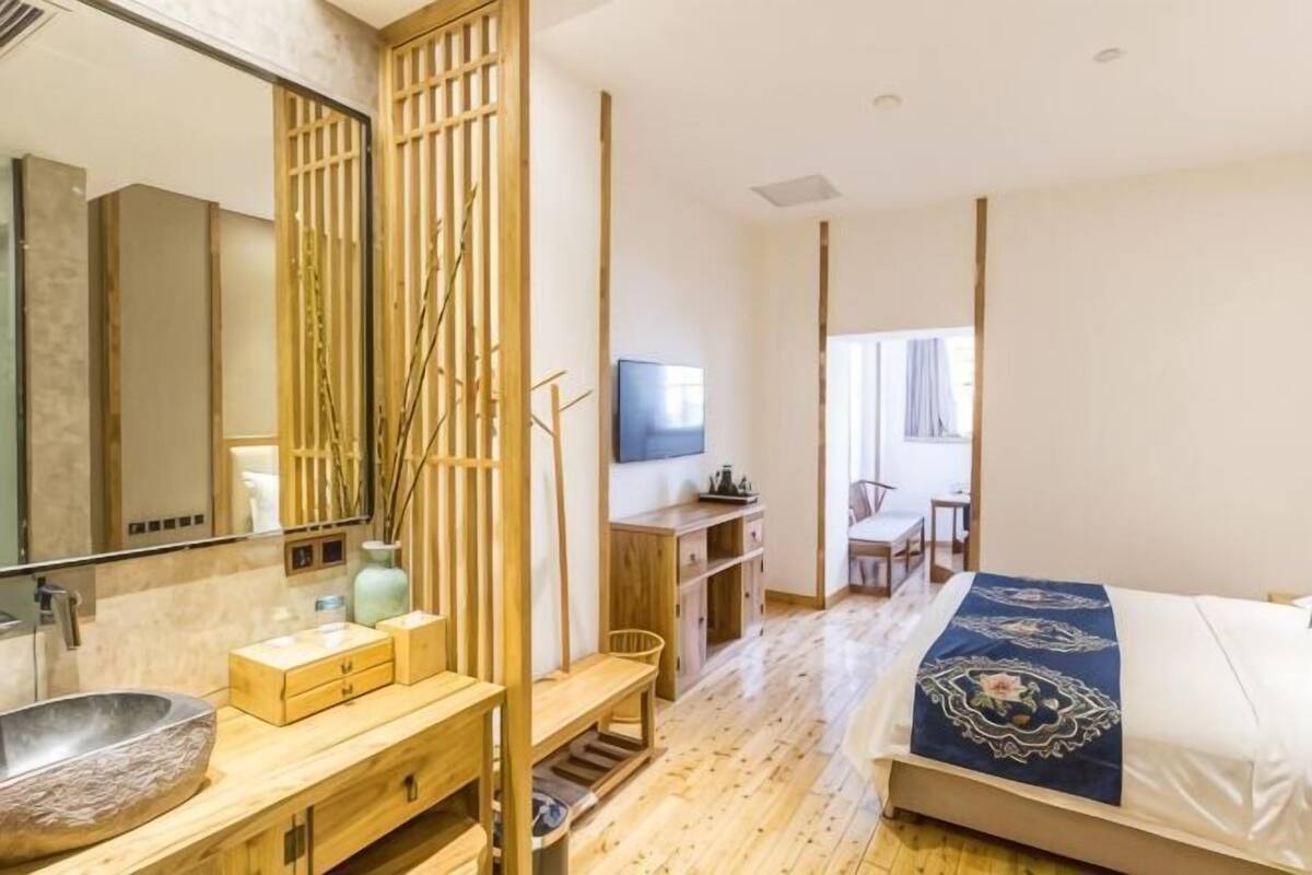 Un dormitorio moderno con suelo de madera, una cama grande con colcha azul, un escritorio de madera, un televisor y un baño con un espejo grande y un lavabo de piedra.