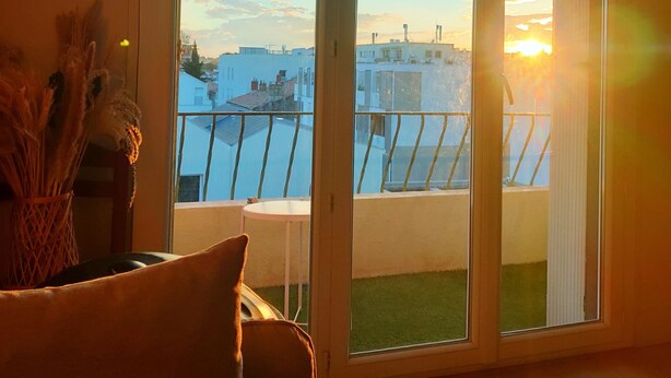 A room with a large sliding glass door leading to a balcony with a view of buildings and a sunset. - South-facing, pleasant, quiet 65 ㎡ (Montpellier)
