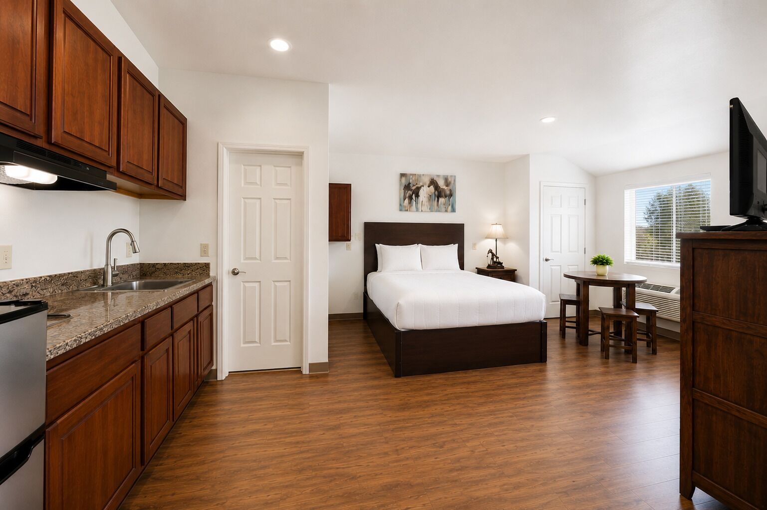 A hotel room with a kitchen area, a bed with white bedding, a small table with a lamp, and a TV on a wooden cabinet.