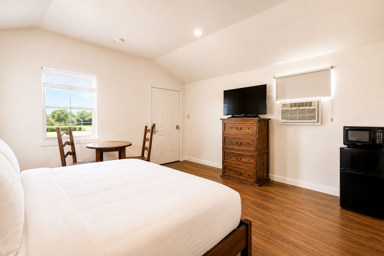 A bedroom with a bed, a dresser, a TV, a microwave, and a window with a view of trees.