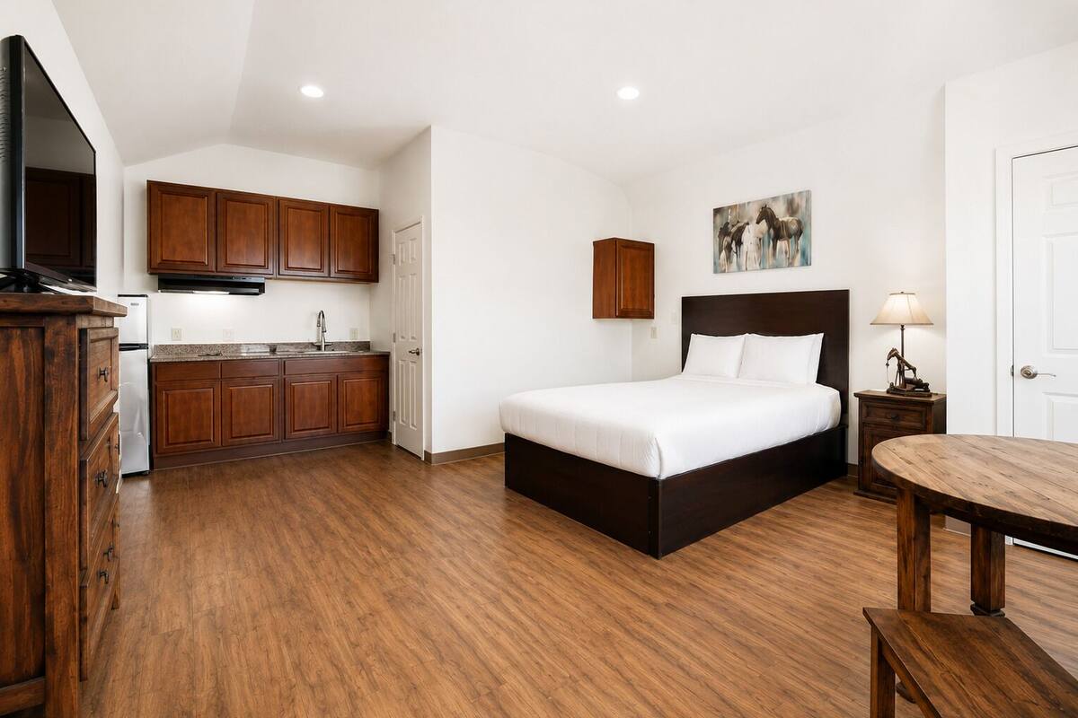 Un dormitorio con una cama, una mesita de noche, una mesa de madera, una zona de cocina con armarios y un cuadro en la pared.