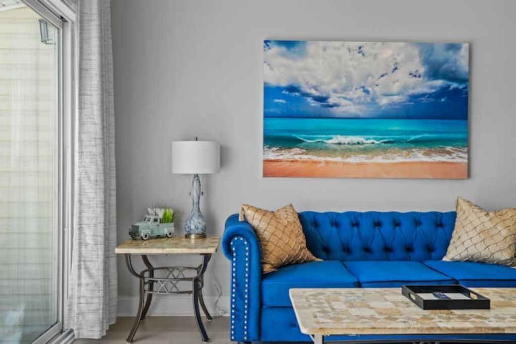 Ein Zimmer mit einem blauen Sofa, einem kleinen Tisch mit einer Lampe und einem Bild von einer Strandansicht.