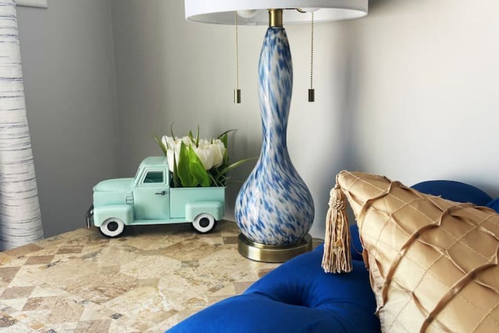 Una habitación con un sofá azul, un camión de juguete turquesa pequeño, una lámpara decorativa y un jarrón con flores blancas sobre un suelo de baldosas.