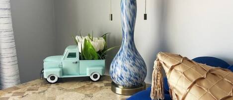 Una habitación con un sofá azul, un camión de juguete turquesa pequeño, una lámpara decorativa y un jarrón con flores blancas sobre un suelo de baldosas.