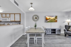 A modern kitchen with white cabinets, a microwave, and a dining area with a wooden table and chairs. A clock and a painting are on the wall.