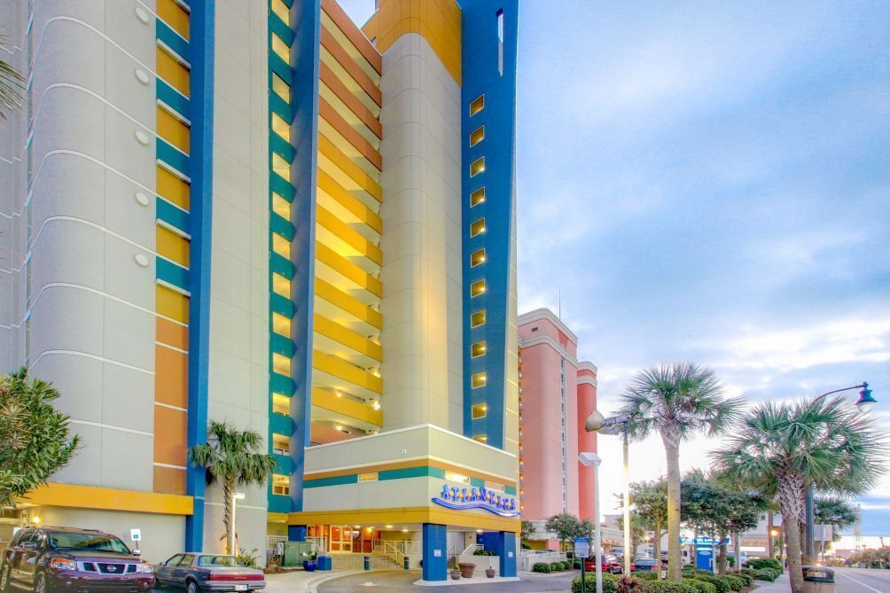A multi-story hotel with a prominent yellow and blue facade, a central entrance, and a palm tree-lined street.