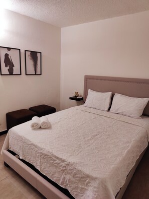 Ein ordentlich bezogenes Bett mit einem gemusterten Bettdeckenüberwurf, zwei Kissen und einem Kopfteil mit Perlendrahtbordüre. An der Wand hängen zwei gerahmte Bilder, ein kleiner Nachttisch mit einer Vase und eine Bank am Fußende des Bettes.