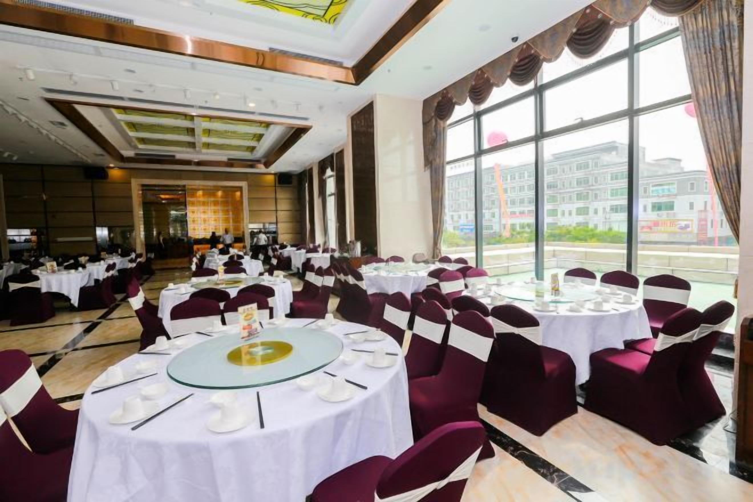 Une salle de banquet avec des tables rondes, des chaises et une vue sur un bâtiment, visible à travers de grandes fenêtres.