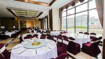 Une salle de banquet avec des tables rondes, des chaises et une vue sur un bâtiment, visible à travers de grandes fenêtres.