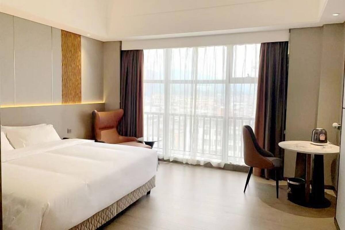 A hotel room with a large bed, a seating area with a table, and a view of the city through large windows.