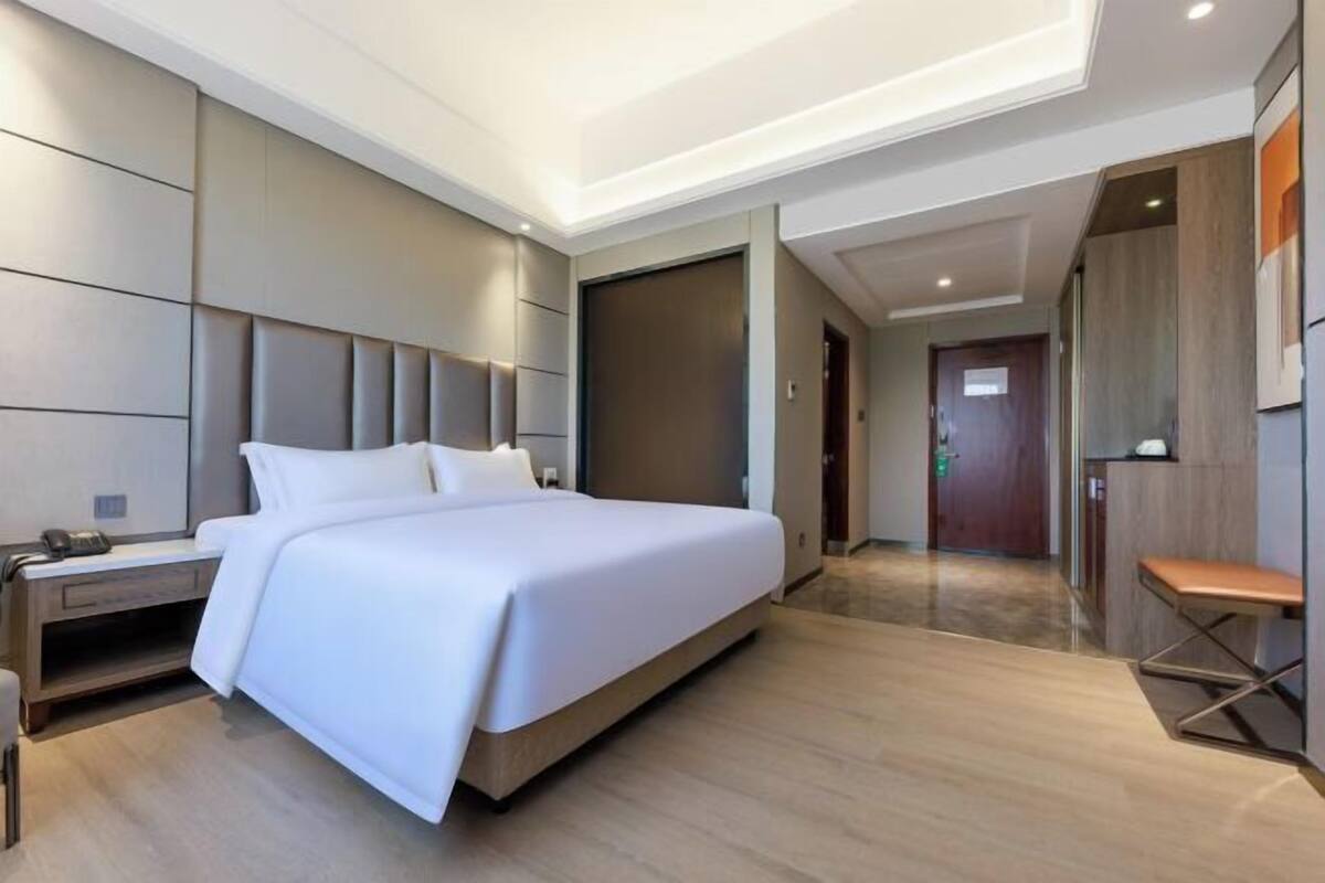 A modern hotel room with a large bed, a bedside table with a phone, a bench, and a door leading to another room.