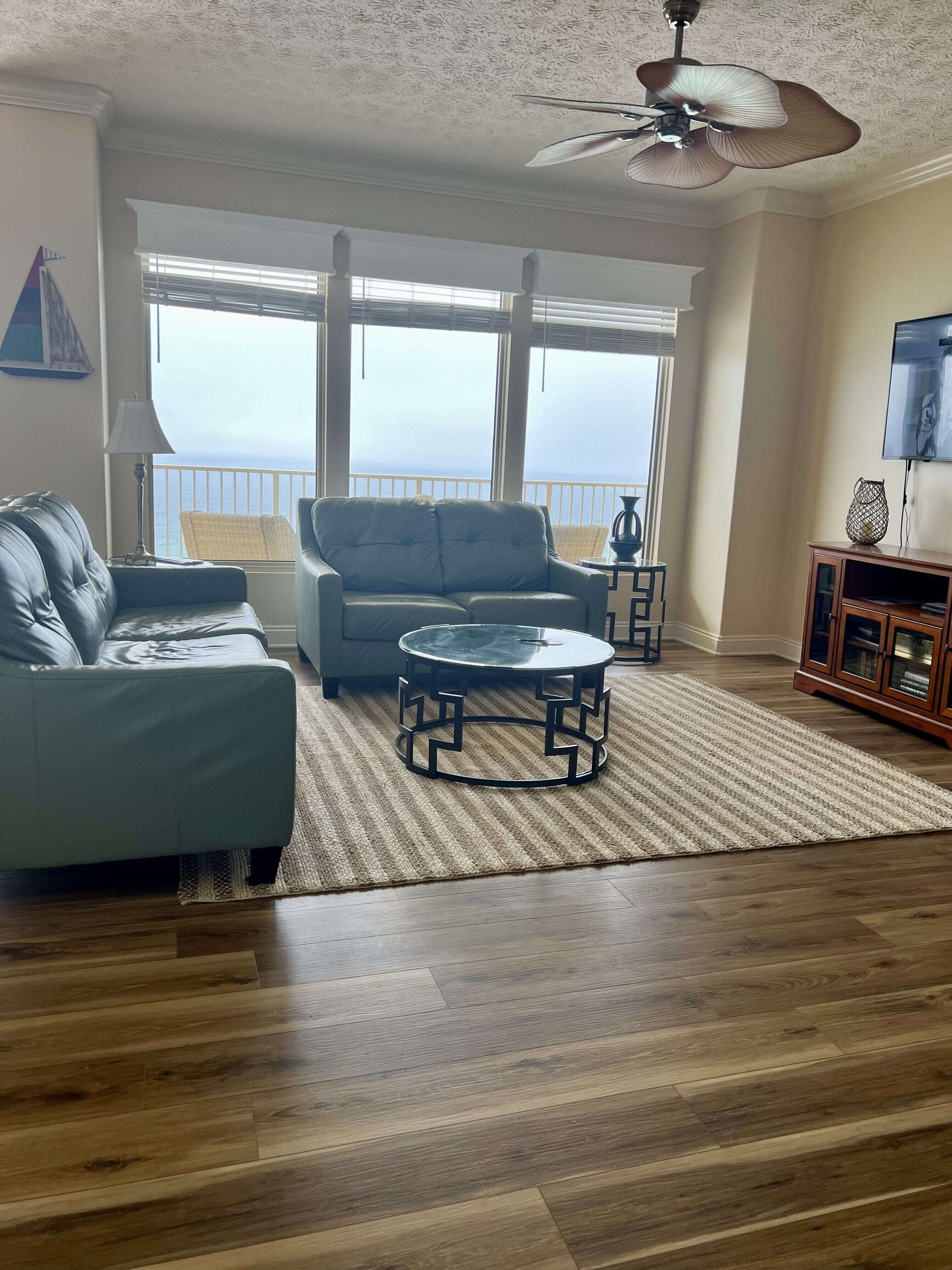 A living room with a sofa, a coffee table, and a TV. There is a balcony with a view of the ocean.