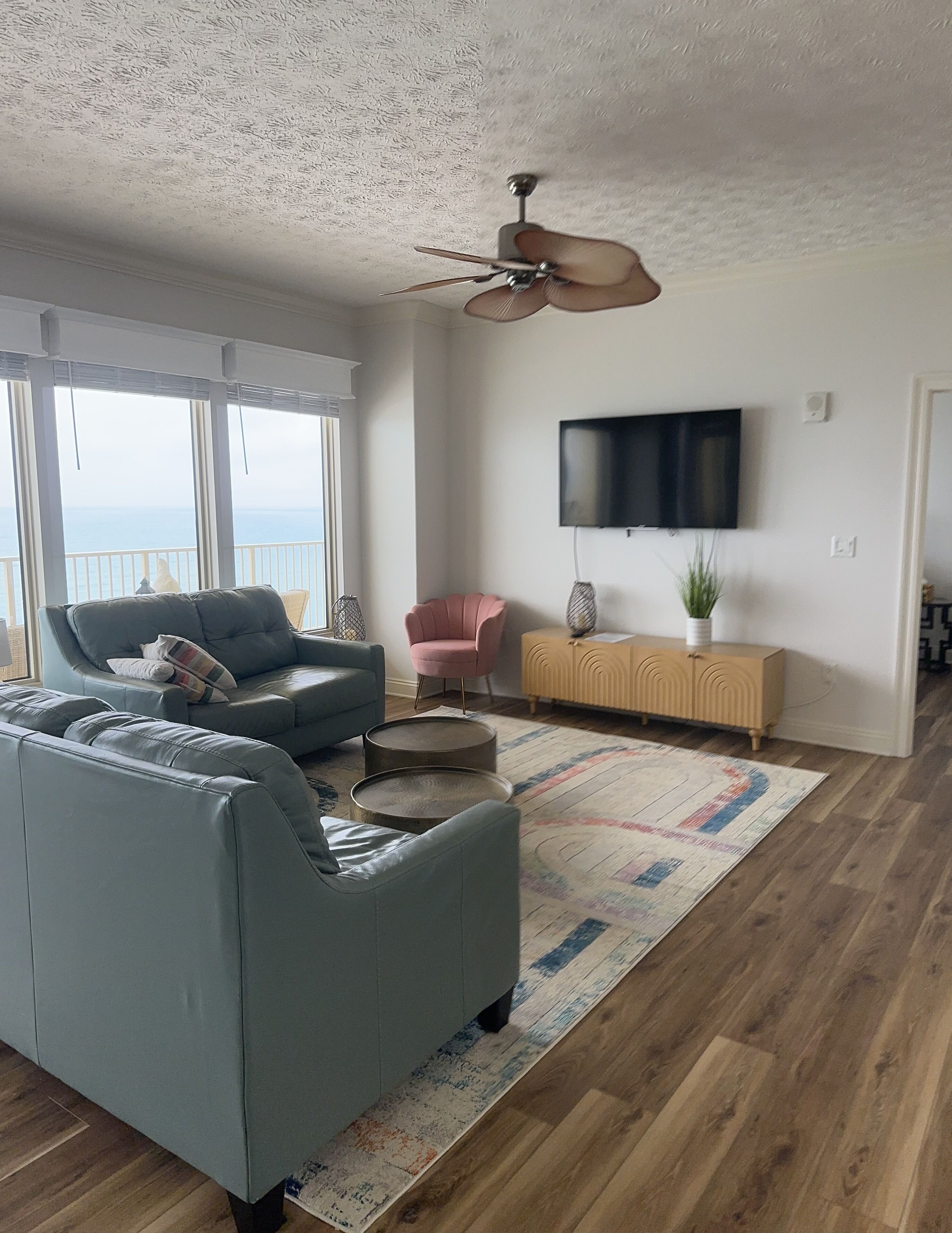 Un salon spacieux avec un canapé gris, un fauteuil rose, un plancher en bois, un ventilateur de plafond et un téléviseur fixé au mur.