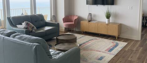 Un salon spacieux avec un canapé gris, un fauteuil rose, un plancher en bois, un ventilateur de plafond et un téléviseur fixé au mur.