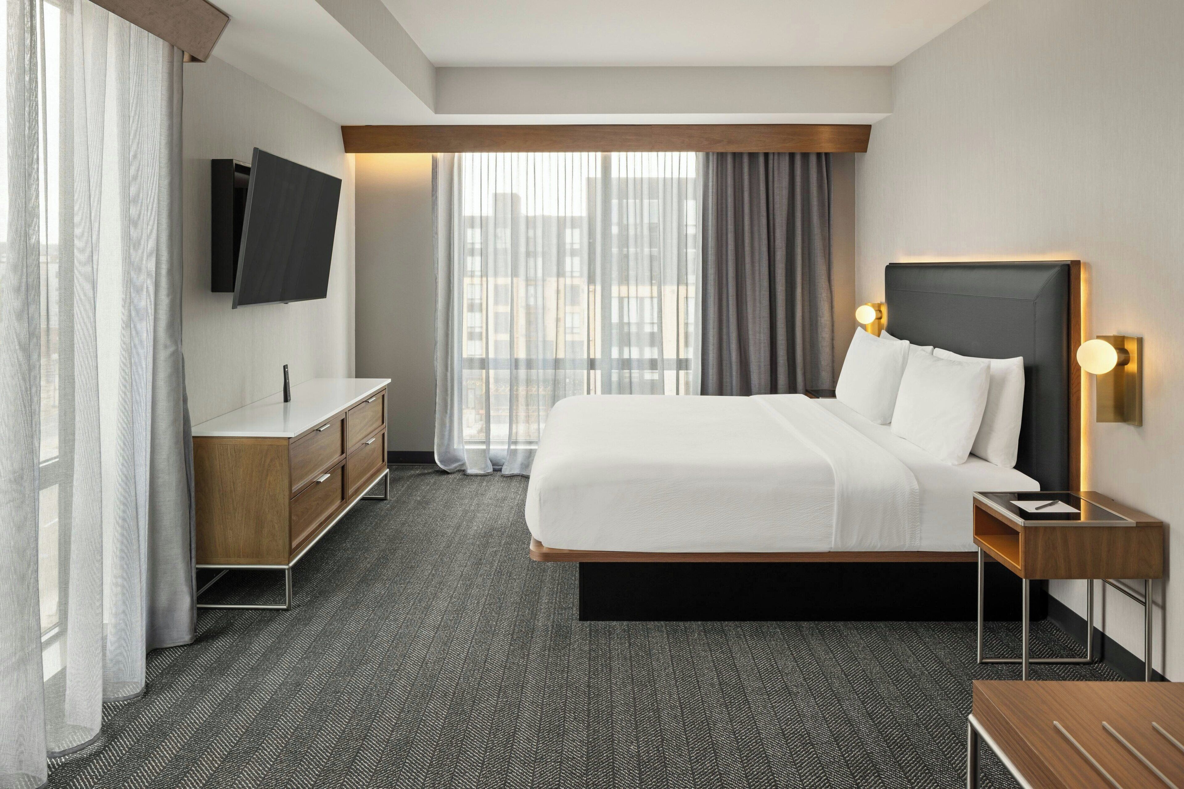 Una habitación de hotel moderna con una cama grande, un televisor de pantalla plana montado en la pared, una mesita de noche de madera y un escritorio con una silla.