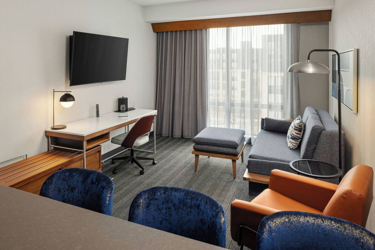 Une chambre d’hôtel équipée d’un téléviseur à écran plat fixé au mur, d’un bureau avec une chaise, d’un canapé avec des coussins, d’un petit pouf et d’une lampe.