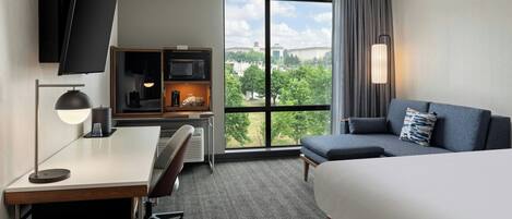 Habitación de hotel con cama, escritorio, silla, televisor y una ventana grande con vistas a la ciudad.