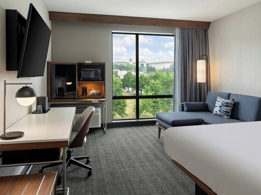 Hotelkamer met een bed, bureau, stoel, televisie en een groot raam met uitzicht op de stad.