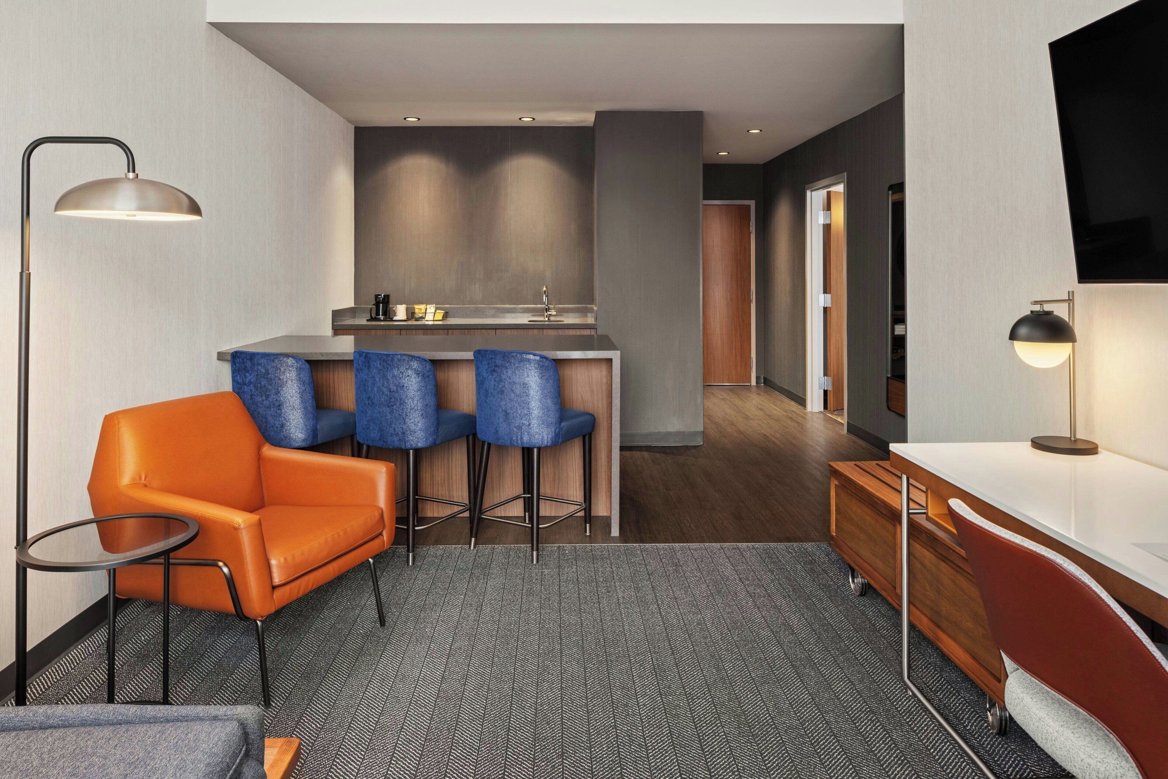 Ein modernes Hotelzimmer mit Küchenzeile, einer Couch, zwei Barhocker, einem Fernseher und einem Schreibtisch.