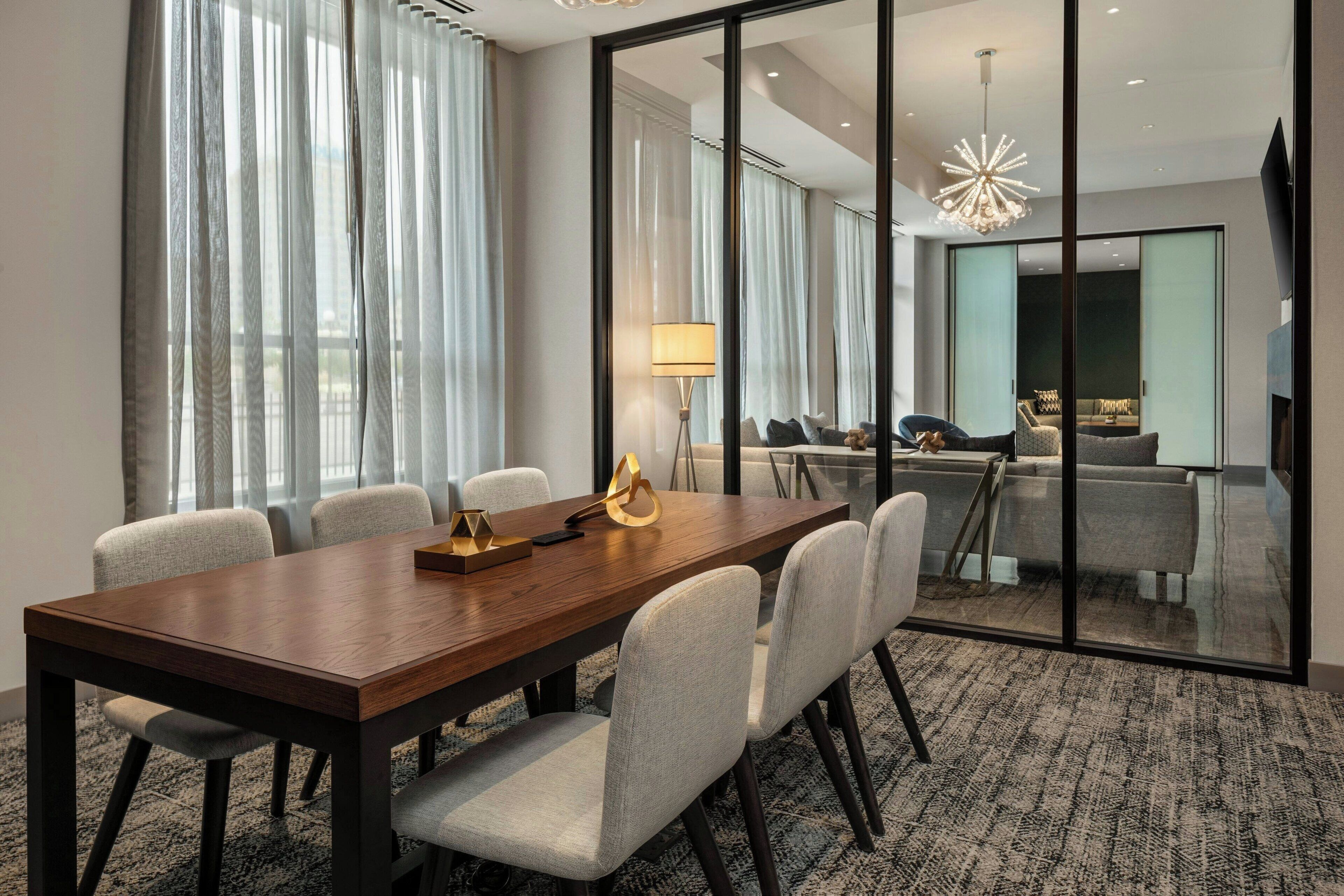 A conference room with a wooden table, grey upholstered chairs, a lamp, and a view through a glass partition to a living area with a sofa and a TV.