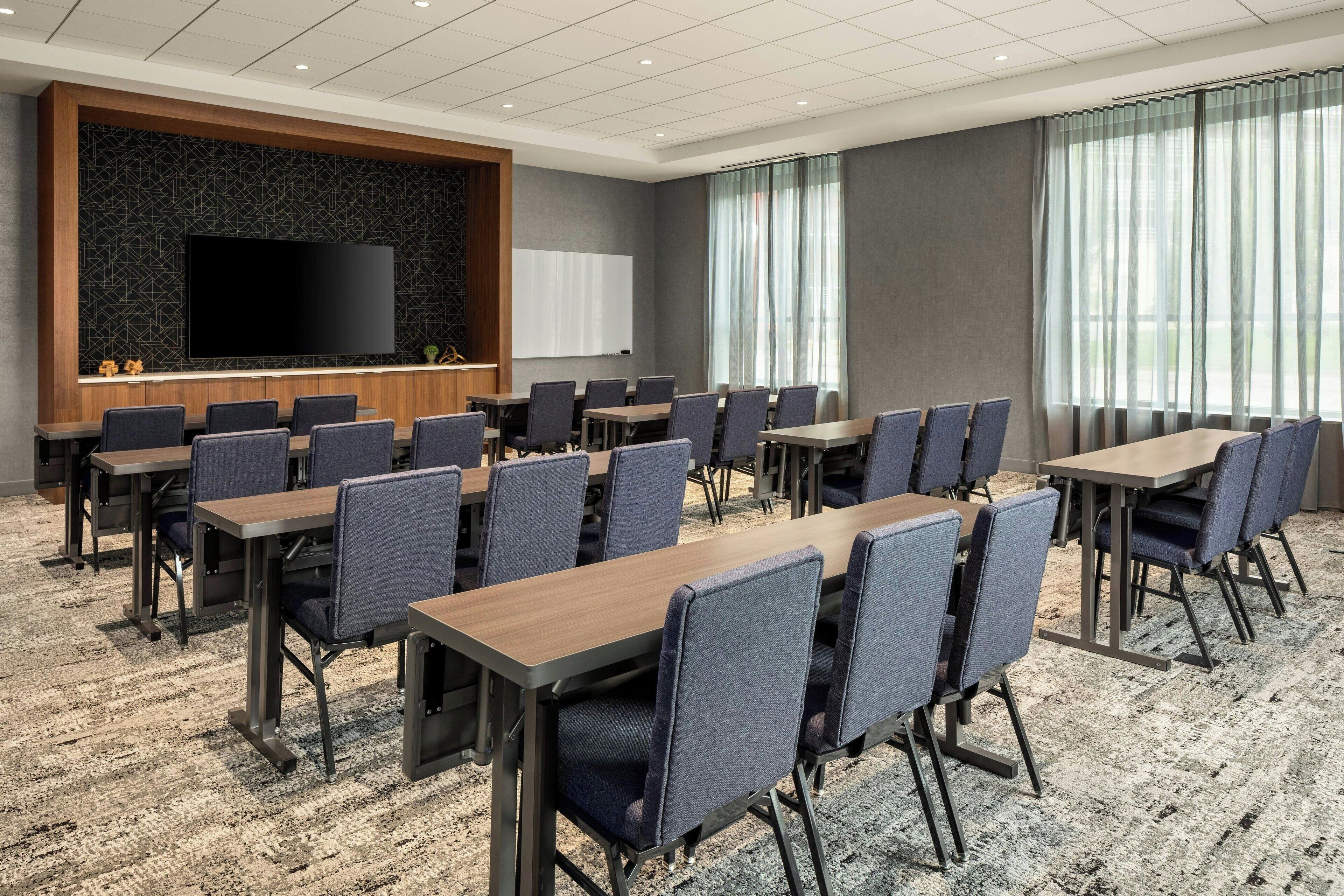 Une salle de conférence équipée d’un grand téléviseur à écran plat, de plusieurs tables et chaises, et dotée d’un sol recouvert de moquette.